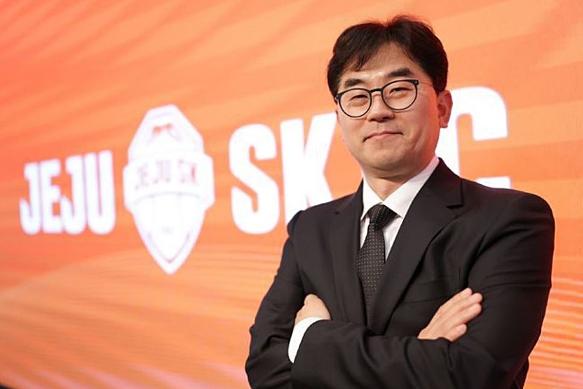 “오렌지색 ‘JEJU SK FC’ 배경과 구단 엠블럼 앞에서, 검은 정장과 넥타이를 착용한 안경 쓴 남성이 팔짱을 낀 채 카메라를 바라보고 있다.”